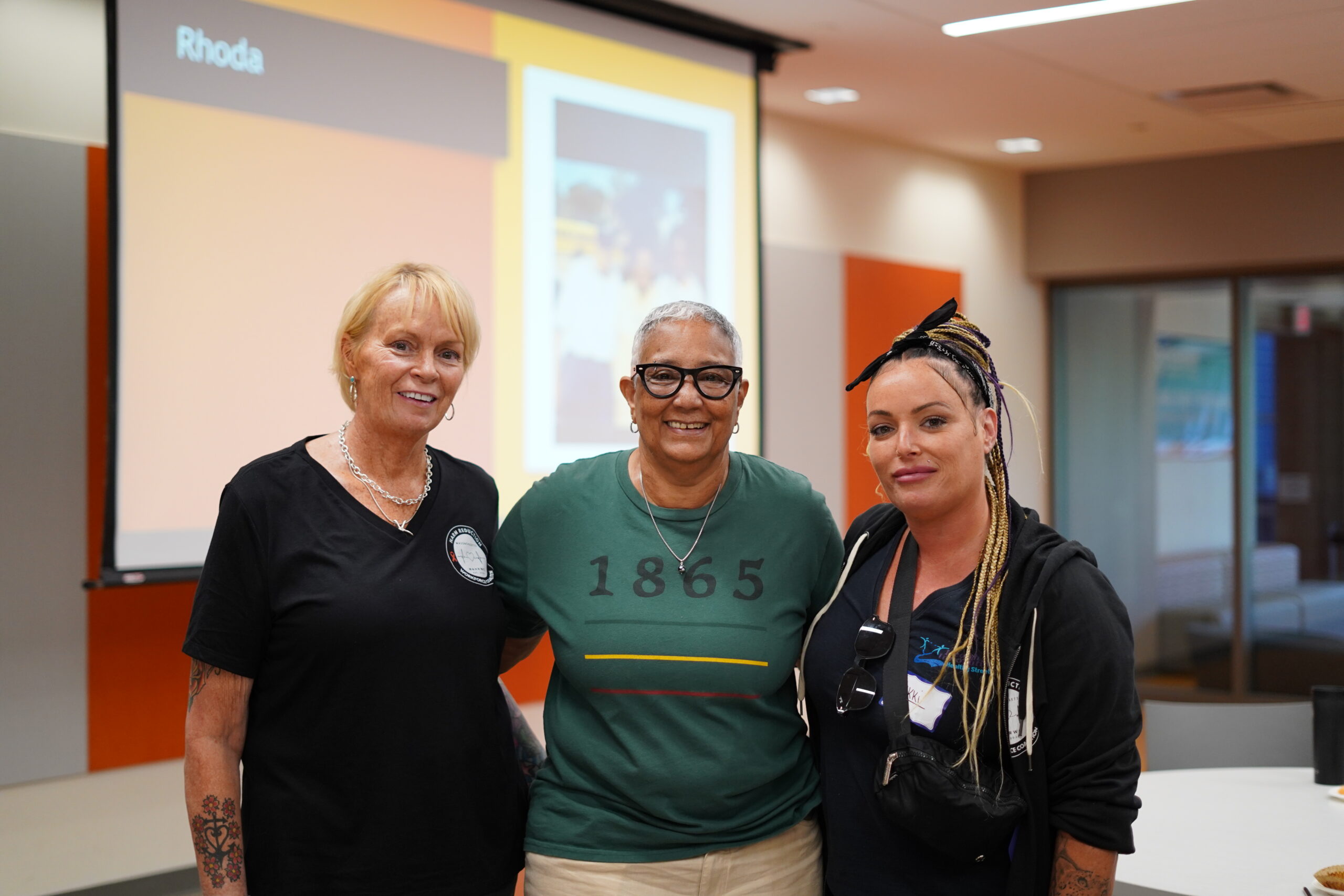 Three women smiling together in front of a presentation screen at a MAHRWC training